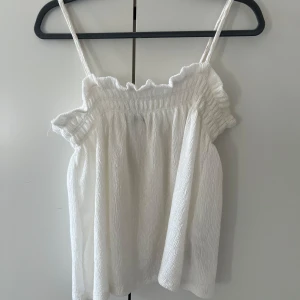 Vit ärmlös topp - Säljer en söt vit ärmlös topp med smala axelband och volangdetaljer vid halsringningen. Perfekt för varma sommardagar. Toppen har en luftig och bekväm passform.