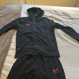 Svart träningsjacka från Under Armour med matchande byxor  - Säljer en svart träningsjacka från Under Armour med dragkedja och huva. Jackan har röda detaljer och logotyp på bröstet. Matchande byxor tillhör.
