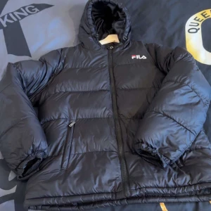 Svart pufferjacka från FILA - Säljer en svart pufferjacka från FILA med huva och dragkedja. Jackan har en stor logga på ryggen och mindre logga på bröstet. Perfekt för kalla dagar med sin vadderade design och långa ärmar.