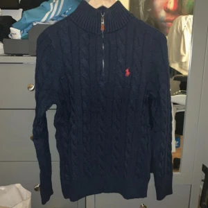 Mörkblå stickad tröja från Polo Ralph Lauren - Säljer en snygg mörkblå stickad tröja från Polo Ralph Lauren med dragkedja vid halsen. Tröjan har ett klassiskt flätmönster och en liten röd logotyp på bröstet. Perfekt för kyligare dagar!