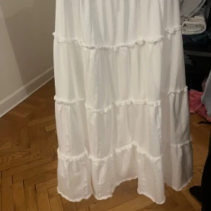 Vit långkjol med volanger - Säljer en vacker vit långkjol med flera lager av volanger. Kjolen har en elastisk midja för bekväm passform och är perfekt för sommarens varma dagar. Den är gjord i ett lätt och luftigt material.