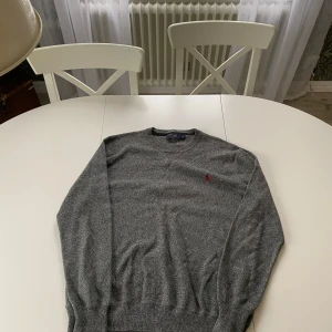 Grå tröja från Polo Ralph Lauren - Säljer en stilren grå tröja från Polo Ralph Lauren med det klassiska röda logotypbroderiet på bröstet. Tröjan har långa ärmar och en rund halsringning, perfekt för en avslappnad look.