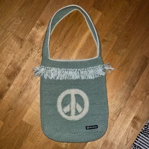 Grön stickad axelväska med fransar - Snygg grön stickad axelväska med ett stort fredsmärke framtill. Väskan har dekorativa fransar och en bred axelrem för bekväm användning. Perfekt för en avslappnad stil.