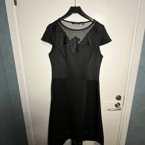 Svart klänning med spetsdetaljer - Elegant svart klänning med korta ärmar och vacker spetsdetalj vid halsringningen. Perfekt för en stilren look.