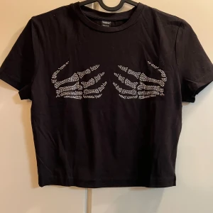 Svart t-shirt med skelettdetaljer från Shein - Cool svart t-shirt från Shein med glittriga skelettdetaljer på framsidan. Perfekt för en edgy look. Kortärmad och croppad design som ger en trendig stil. Tillverkad i mjukt material för bekväm passform.
