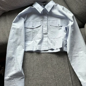 Ljusblå croppad skjorta från H&M Divided - Säljer en stilren ljusblå croppad skjorta från H&M Divided. Skjortan har långa ärmar och två bröstfickor med knappar. Perfekt för en trendig look!