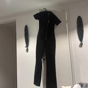 Svart jumpsuit med dragkedja - Snygg svart jumpsuit med dragkedja framtill. Perfekt för en stilren look, oavsett om det är vardag eller fest. Den har en smickrande passform och är superbekväm att bära. Ett måste i garderoben för alla som älskar enkel elegans! Köpt på Plick och endast använd en gång av mig! Säljer då den är för stor!