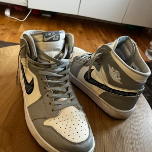 Grå och vita sneakers från Dior x Nike - Snygga grå och vita sneakers från samarbetet mellan Dior och Nike. Skorna har en hög modell med snörning och den ikoniska Nike-loggan i ett unikt mönster. Perfekta för den som vill ha något exklusivt och stilrent.