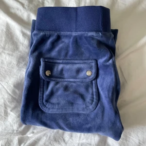 Blå juicy byxor med fickor - Säljer ett par bekväma marinblå juicy byxor med bred resår i midjan och praktiska fickor med knappar. Byxorna har en loose passform och är perfekta för en avslappnad stil. Har dock ett litet hål precis nedanför höger ficka.💙