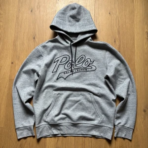 Grå hoodie från Ralph Lauren - Säljer en snygg grå hoodie från Ralph Lauren med stor logga på bröstet. Tröjan har en klassisk känguruficka och justerbar huva med dragsko. Perfekt för en avslappnad stil.