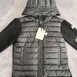 Moncler cardigan  - Snygg svart pufferjacka från Moncler med huva och dragkedja. Jackan har långa ärmar i ett stickat material och en praktisk ficka med dragkedja på ärmen. Perfekt för kyliga dagar!