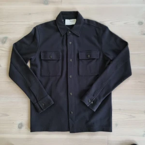 Svart overshirt från Selected Homme - Stilren svart overshirt från Selected Homme med klassisk krage och två stora bröstfickor med knappar. Skjortan har långa ärmar och knäppning framtill, perfekt att bära som lager över en t-shirt. Materialet känns mjukt och lite tjockare. Skriv ifall du har fler funderingar.