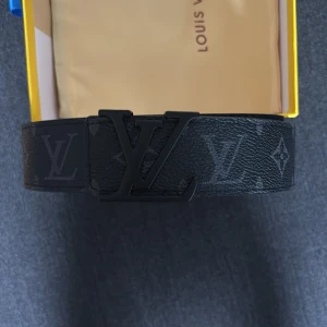 Svart Louis Vuitton monogram bälte - Snyggt svart bälte från Louis Vuitton med klassiskt monogrammönster och stor LV-logga i matt svart metall som spänne. Bältet är gjort i präglat skinn och har en stilren, modern vibe som lyfter vilken outfit som helst.