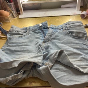 Ljusblå jeansshorts i denim - Säljer ett par ljusblå jeansshorts i klassisk denim med fyra fickor, bälteshällor och råa benslut. Shortsen har dragkedja och knapp framtill och är perfekta för varma dagar. Passar dig som gillar en avslappnad och tidlös stil.