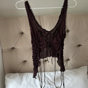 Brun croppad snörad linnetopp - Säljer en brun croppad linnetopp med snörning framtill och rynkade detaljer längs sidorna. Toppen har spetskantade axelband och är ärmlös, med transparent meshmaterial som ger en edgy vibe. Perfekt för dig som gillar unika och trendiga plagg.