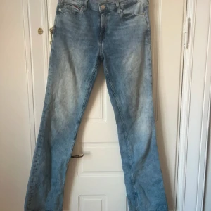 Tommy Jeans bootcut ljusblå jeans - Säljer ett par ljusblå jeans från Tommy Jeans i modellen Mid Rise Boot. Jeansen har klassisk femficksdesign, raka ben med bootcut och snygg tvätt. Tillverkade i bomull för en skön känsla och avslappnad look.