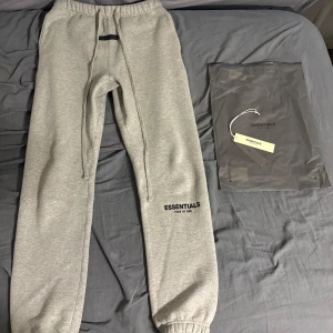 Grå Essentials Fear of God mjukisbyxor - Snygga grå mjukisbyxor från Essentials Fear of God med broderad logga på benet. Byxorna har elastisk midja med dragsko, muddar vid benslut och är gjorda i mjuk bomullsblandning. Perfekta för chill eller streetwear vibes.