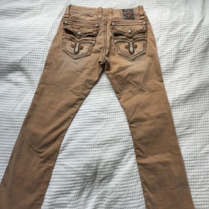 Bruna byxor med broderade bakfickor - Snygga bruna byxor från Rock Revival med detaljerade broderier och knappar på bakfickorna. Byxorna har raka ben, coola kontrastsömmar och unika detaljer i orange och svart på insidan av linningen. Perfekta för dig som gillar statement-plagg.