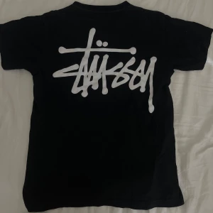 Svart Stüssy t-shirt med logga - Svart t-shirt från Stüssy med stor vit graffiti-inspirerad logga på ryggen och mindre logga på bröstet. Klassisk rund hals och korta ärmar. Skön passform och tillverkad i mjuk bomull, perfekt för streetwear-stilen.
