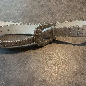Vitt skärp med glittrigt spänne - Säljer ett vitt bälte med glittrande detaljer och ett stort, dekorerat spänne i silver med strass och kors. Bältet har silverfärgade nitar och pärlor längs kanterna som ger en extra bling-effekt. Aldrig använt