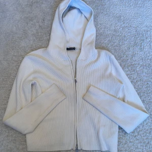 Kofta från Brandy Melville - Säljer en vit ribbad hoodie från Brandy Melville med dragkedja framtill och huva. Tröjan har långa ärmar och är gjord i ett mjukt stickat material som är perfekt för chill dagar. Enkel och clean design som passar till allt.