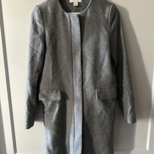 Grå kappa från H&M, storlek 34 - Stilren grå kappa från H&M i storlek 34. Rak modell med dold dragkedja framtill och två fickor med lock. Kappan har rundad halsringning och är helfodrad, perfekt för kyligare dagar.