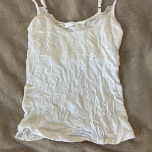 Vit spetslinnetopp från Stories XS - Supersöt vit linnetopp från Stories i storlek XS. Toppen har tunna axelband och en fin spetskant upptill som ger en romantisk vibe. Perfekt att styla under en skjorta eller bara som den är under varma dagar.