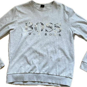 Grå sweatshirt från Hugo Boss - Snygg ljusgrå sweatshirt från Hugo Boss med stort BOSS-tryck i svart, vitt och svart på bröstet. Tröjan har rund hals, långa ärmar och ribbade muddar. Perfekt för en chill och stilren look.