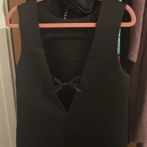 Svart kort klänning från Divided - Supersnygg svart kort klänning från Divided med djup v-ringning och en söt rosettdetalj framtill. Ärmlös modell med enkel, stilren design som passar perfekt till sneakers eller boots. Gjord i ett mjukt polyestertyg som känns skönt mot huden. Använd en gång ❣️