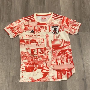 Japan Tokyo fotbollströja Adidas - Unik Japan fotbollströja från Adidas med Tokyo-inspirerat rött och vitt mönster över hela tröjan. Klassiska tre ränder på axlarna, Japan-märke på bröstet och coola japanska motiv. Tillverkad i lätt och ventilerande material, perfekt för match eller träning.