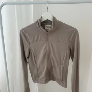 Beige träningsjacka från H&M Move - Snygg beige träningsjacka från H&M Move med dragkedja framtill och hög krage. Jackan har en clean look, långa ärmar och smidig passform. Perfekt för dig som gillar att röra på dig och vill ha en stilren jacka till gymmet eller promenaden. I nyskick, endast testad.