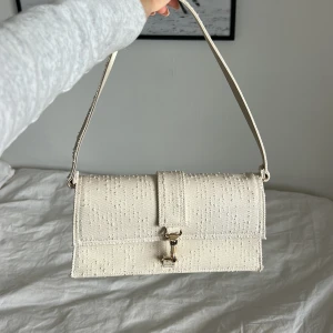 Beige axelväska i textil med spänne från hm - Snygg beige axelväska i textil med strukturerad yta och guldfärgat spänne framtill. Väskan har en rektangulär form och justerbar axelrem, perfekt för att bära det viktigaste. Enkel men trendig design som passar till många outfits.