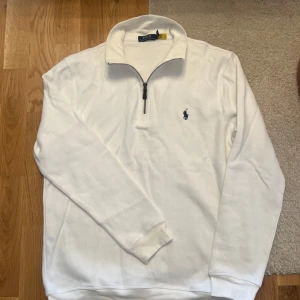 Polo Ralph Lauren Halfzip - Vit långärmad tröja från Polo Ralph Lauren med klassisk half zip-dragkedja och broderad marinblå logga på bröstet. Tröjan har hög krage och är gjord i mjuk bomull, perfekt för en clean och stilren look.