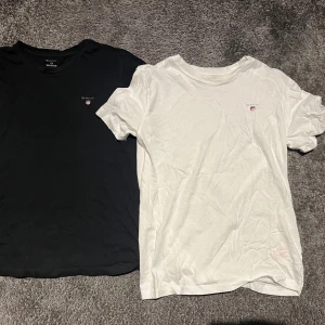 Svart och vit t-shirt från Gant - Två klassiska t-shirts från Gant, en svart och en vit, båda med liten broderad logotyp på bröstet. T-shirtsen har rund hals och korta ärmar, tillverkade i mjuk bomull för en skön känsla. Perfekta för en clean och enkel stil. passar mig som är 179 cm. Vita är helt oanvänd