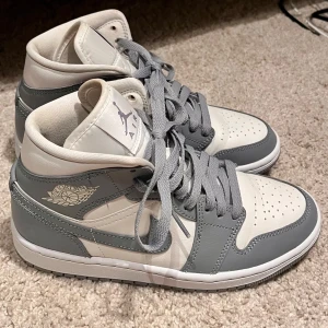 Nike Air Jordan 1 Mid grå/vit - Snygga Nike Air Jordan 1 Mid sneakers i grått och vitt skinn. Endast använt dom 1 gång och då endast en liten stund inomhus. Klassisk siluett med grå snörning, perforerad tå och ikoniska Air Jordan-loggan på sidan. 