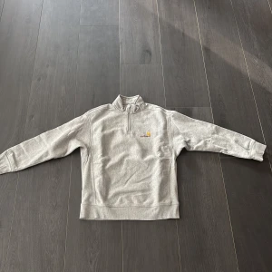 Grå half zip tröja från Carhartt - Säljer en grå långärmad tröja från Carhartt med broderad logga på bröstet. Tröjan har en hög krage och half zip-dragkedja framtill. Mjuk insida och klassisk passform, perfekt för lager-på-lager under kyliga dagar.