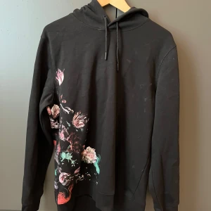 Svart hoodie med blommigt tryck H&M - Svart hoodie från H&M i storlek M med snyggt blommigt tryck i rosa, rött och grönt på både fram- och baksidan. Tröjan har huva med snörning och långa ärmar. Perfekt för dig som gillar streetstyle med en twist av färg.
