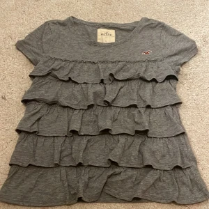 Grå volangblus från Hollister - Supersöt grå blus från Hollister med flera lager volanger framtill. Den är storlek S men sitter som XS.  Perfekt för dig som gillar en chill men ändå söt stil.