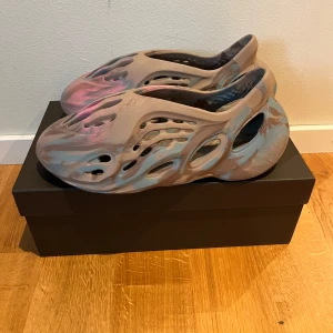 Adidas Yeezy Foam Runner sand/blå - Unika Adidas Yeezy Foam Runner med futuristisk design i sandfärgad bas med blå och rosa detaljer. Skorna är tillverkade i lätt syntetmaterial med många hål för maximal ventilation och en chunky, vågig sula. Perfekta för dig som vill sticka ut med en modern vibe.
