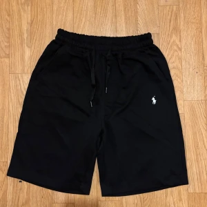 Svarta shorts från Polo Ralph Lauren - Svarta shorts från Polo Ralph Lauren med klassisk broderad logga i vitt på vänster ben. De har elastisk midja med snörning och är tillverkade i mjuk bomull. Passformen är loose och de har två sidofickor. Perfekta för chill dagar eller sport.