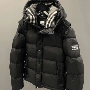Svart pufferjacka från Burberry - Snygg svart pufferjacka från Burberry med klassisk logga på ärmen och rutigt foder i huvan. Jackan har dragkedja och knappar framtill samt en mysig, vadderad känsla som håller dig varm. Perfekt för kalla dagar och riktigt stilren look.