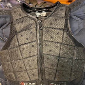 Svart skyddsväst från Racesafe barn M - Skyddsväst från Racesafe i svart med quiltad design och dragkedja framtill. Västen är tillverkad i syntetmaterial och har ventilerande meshpaneler. Perfekt för ridning och ger bra rörelsefrihet tack vare den flexibla konstruktionen.