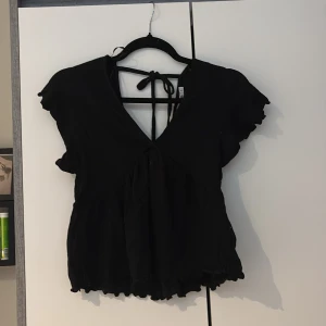 Svart blus med knyt från 157 - Svart blus från 157 med v-ringning och knyt i ryggen. Modellen har korta, volangiga ärmar och lätt rynkad nederkant. Tunt, flowy tyg som ger en luftig känsla. Perfekt för dig som gillar en feminin och avslappnad stil.