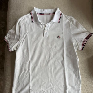 Vit Moncler piké med röd detalj - Vit pikétröja från Moncler med klassisk krage och korta ärmar. Tröjan har Moncler-logga broderad på bröstet och snygga röd-svarta ränder på krage och ärmslut. Tillverkad i mjuk bomull som känns skön mot huden. Perfekt för dig som gillar stilrena märkesplagg. Nypris 3500