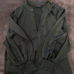 Mörkgrön från Anna Field - Supersnygg mörkgrön från Anna Field med v-ringning och långa puffärmar. Ärmsluten har breda smockdetaljer med volangkant.har en mjuk, glansig yta som ger en lyxig känsla.