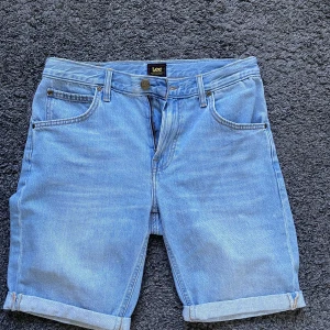 Ljusblå jeansshorts från Lee - Snygga ljusblå jeansshorts från Lee med klassisk femficksdesign och uppvikta benslut. Shortsen har normal passform, bälteshällor och dragkedja framtill. Perfekta för varma dagar och enkel att matcha med allt.