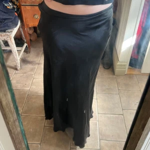 Svart långkjol i satin från 157 Collection - Säljer en svart långkjol i glansigt satinmaterial från 157 Collection. Kjolen har en enkel och stilren design med lätt utsvängd passform som ger snyggt fall. Perfekt att styla med crop top eller stickad tröja för en trendig look.