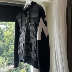 Svart Moncler hoodie med vadderad front - Svart hoodie från Moncler med vadderad, quiltad front i syntet och stickade ärmar. Stor vit M-detalj på ena ärmen och klassisk Moncler-logga. Huva med snörning och dragkedja framtill. Perfekt för kyliga dagar och riktigt snygg streetstil.