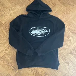 Svart hoodie med tryck från corteiz  - Svart hoodie från Corteiz i storlek medium med ett vitt tryck av en byggnad på bröstet. Tröjan har huva och långa ärmar, perfekt för dig som gillar streetwear och vill ha en chill look. Materialet känns mjukt och bekvämt, troligen bomull eller polyester. Tröjan är M dock stor så L 