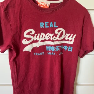 Vinröd Superdry t-shirt med tryck - Snygg vinröd t-shirt från Superdry med stort vitt och blått tryck på bröstet. Klassisk rund halsringning och korta ärmar. Skön bomullskvalitet och avslappnad passform, perfekt till jeans eller shorts.